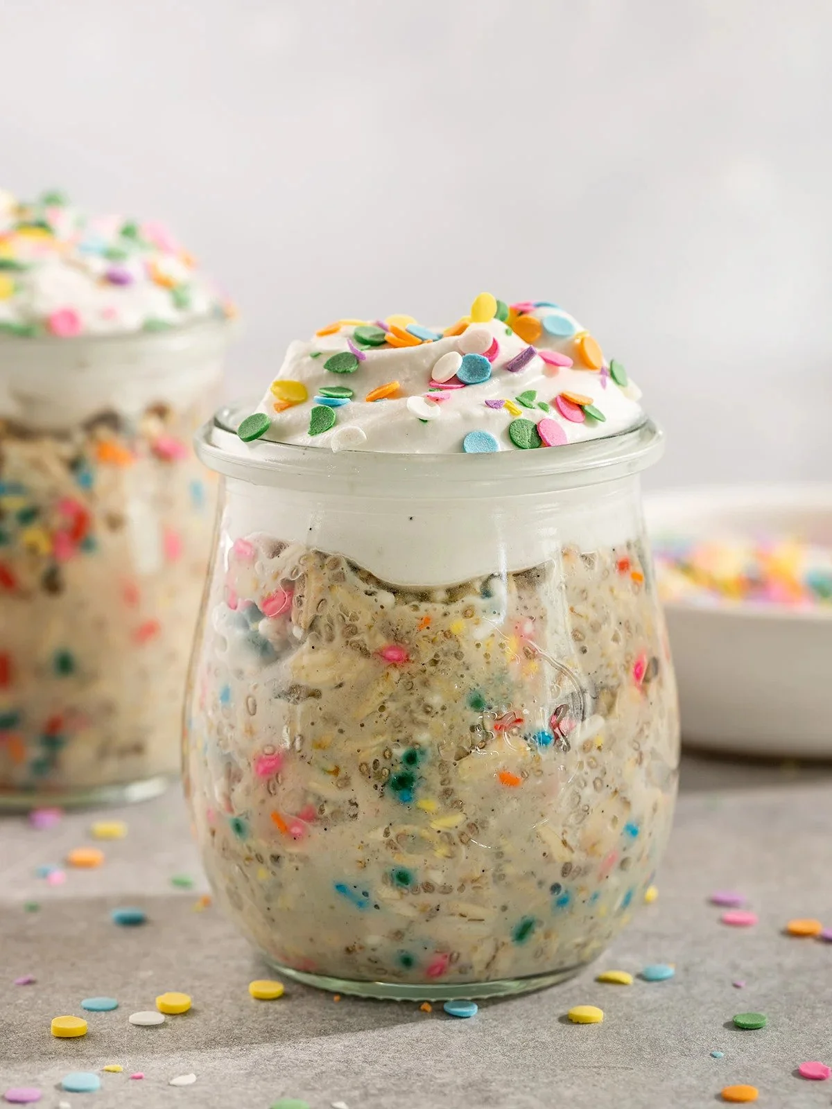 A glass jar filled with vegan cookie mixture topped with whipped cream and colorful sprinkles, with more sprinkles scattered around on a surface.