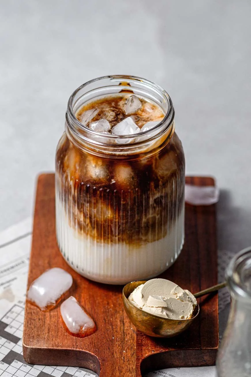A glass jar of iced coffee with milk on a wooden board, with ice cubes inside the drink and outside on the board. There is a spoon with vanilla ice cream next to the jar. The background is a light surface.