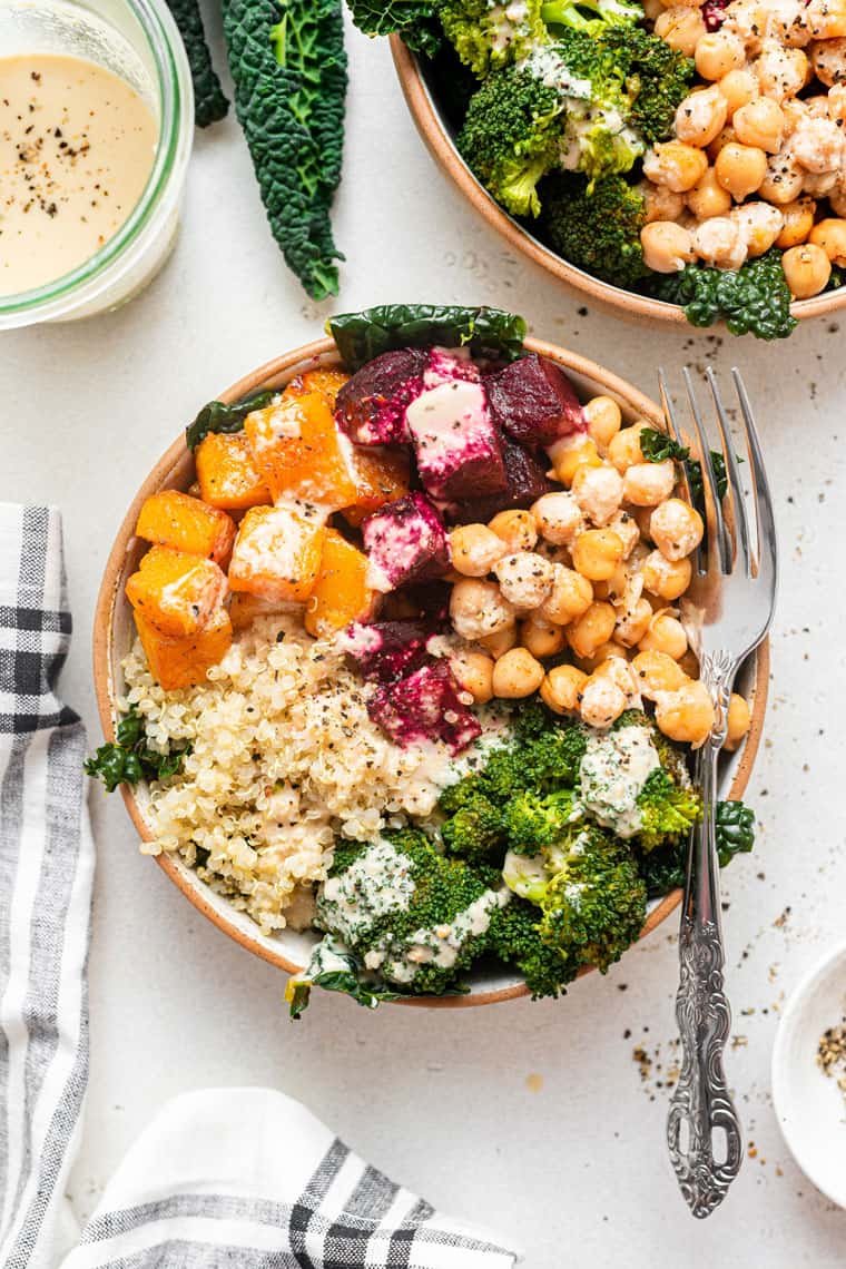 A bowl with roasted butternut squash, beets, cauliflower rice, broccoli, and chickpeas, served with a side of tahini sauce and fresh kale leaves.
