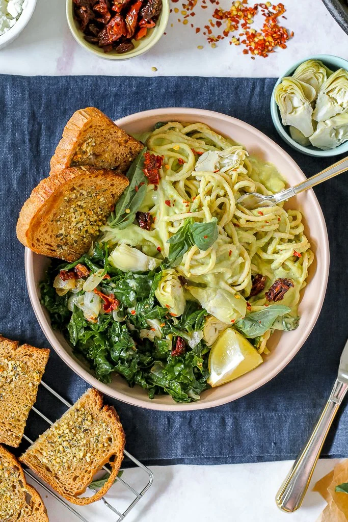 A bowl of pasta with sliced artichokes, kale, and red pepper flakes, served with lemon wedges and garlic bread on the side.