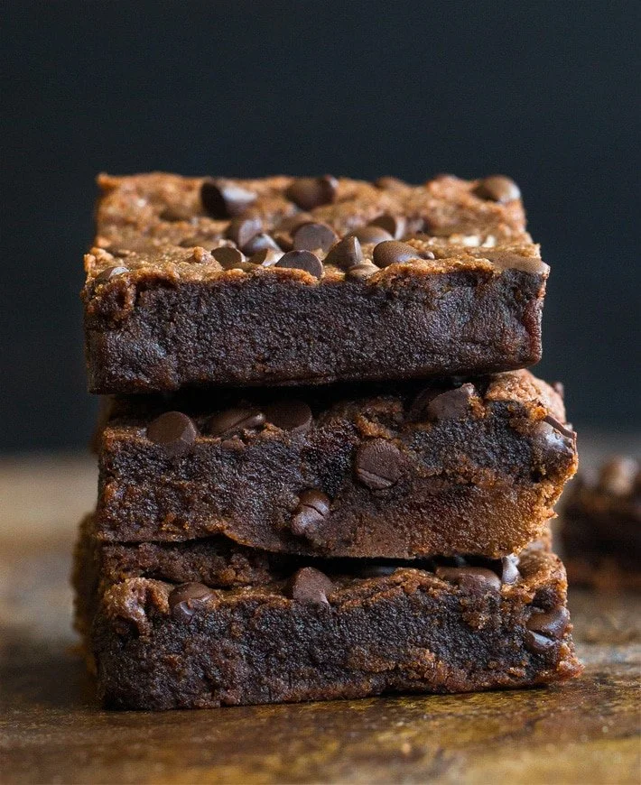 Stack of three chocolate brownie bars topped with chocolate chips on a wooden surface with a dark background.