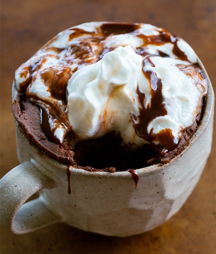 A ceramic mug filled with hot chocolate topped with whipped cream and drizzled with chocolate syrup, placed on a wooden surface.