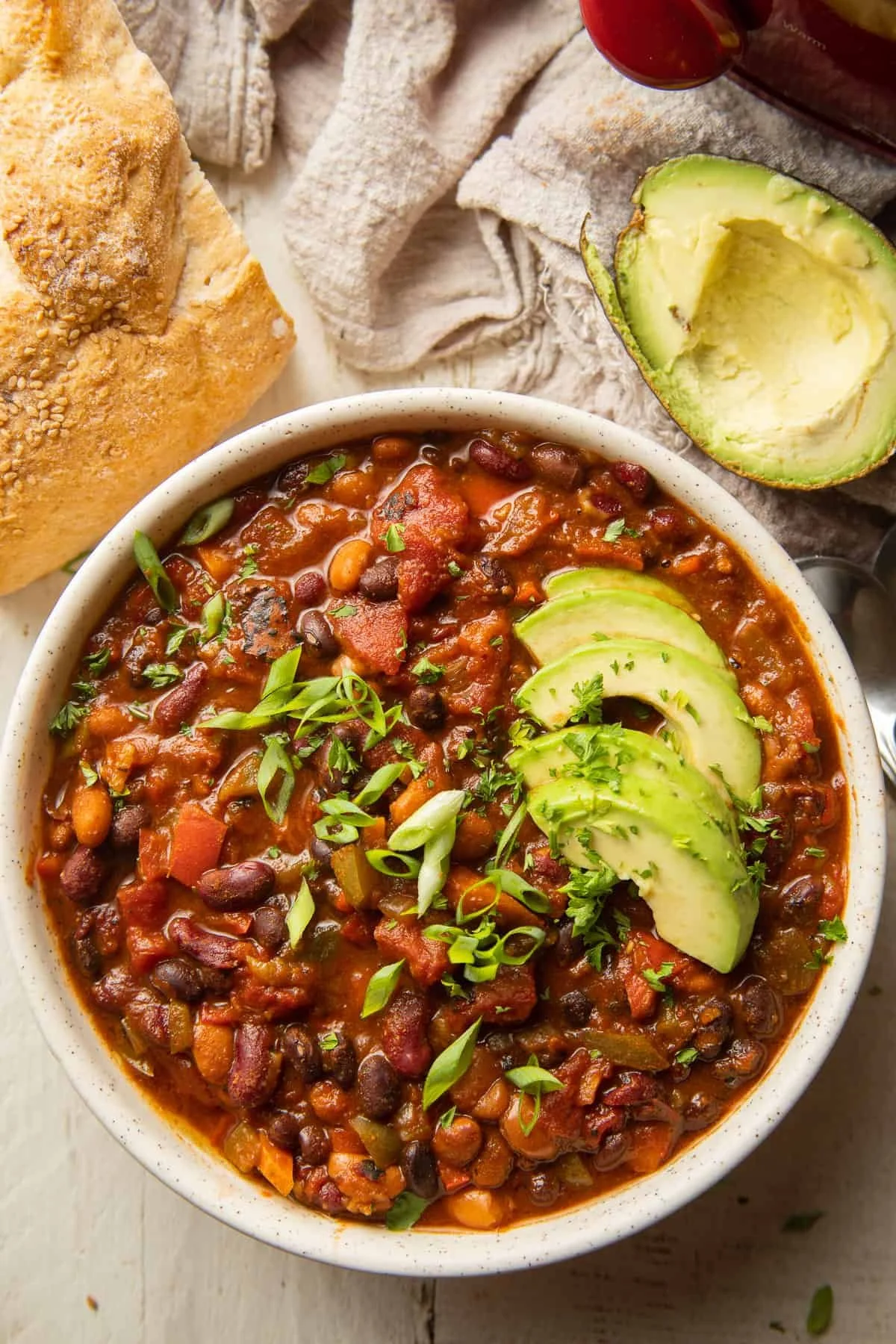 Bowl of chili with black beans, kidney beans, and chopped vegetables garnished with sliced avocado and green onions, next to bread and halved avocado on a cloth.