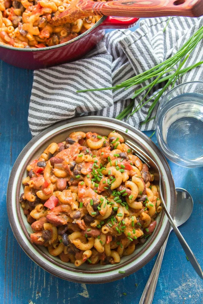 A bowl of pasta salad with macaroni, black beans, chopped tomatoes, and chopped green onions on a blue table, with a glass of water nearby, and a striped cloth and green onions in the background.
