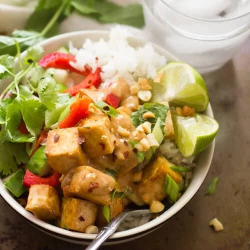 Bowl of salad with tofu, rice, corn, red pepper, cucumber, cilantro, and lime wedges, with a glass of water in the background.