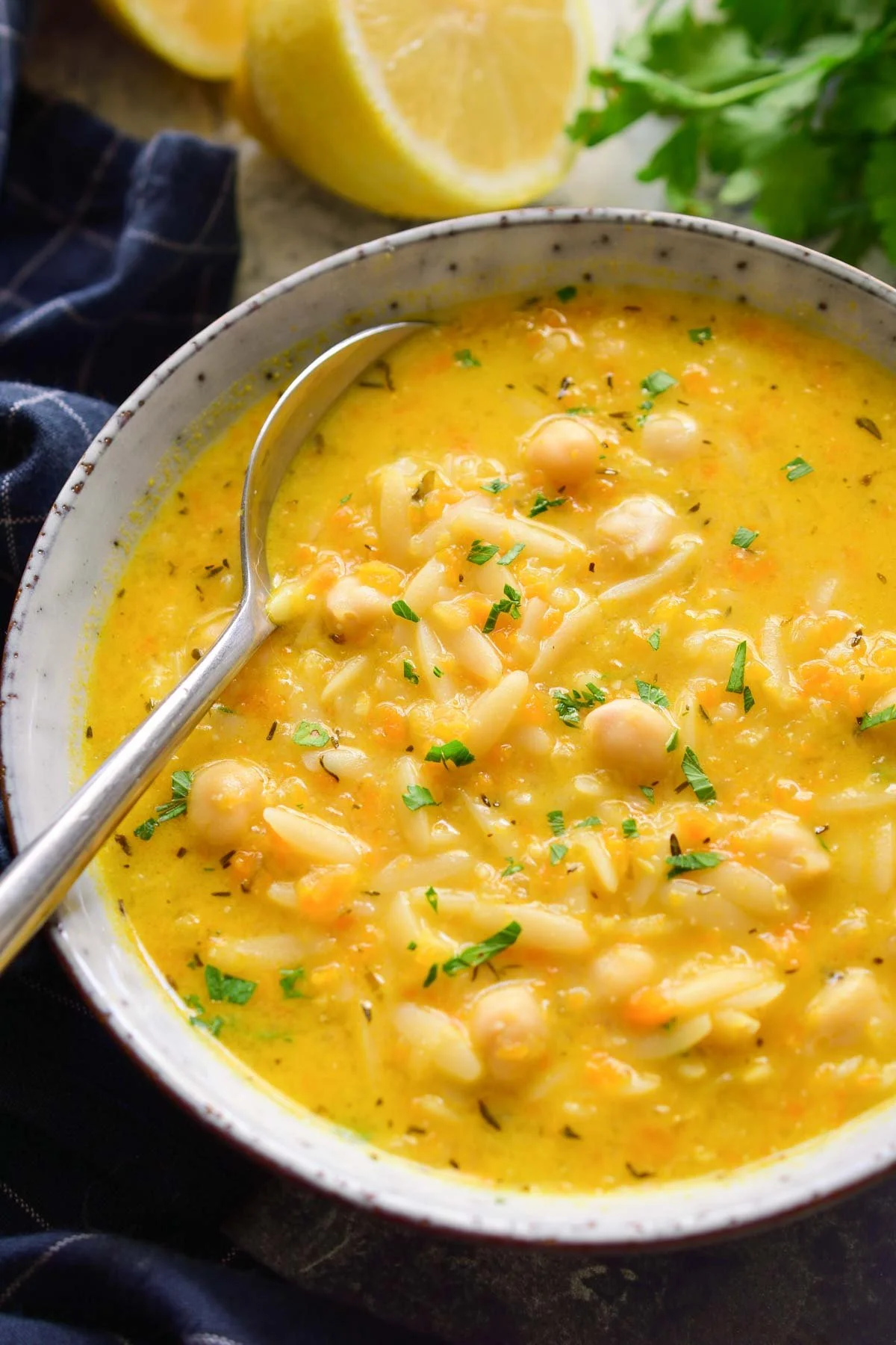 A bowl of creamy vegan orzo chickpea soup garnished with chopped herbs, with lemon halves and fresh parsley in the background.