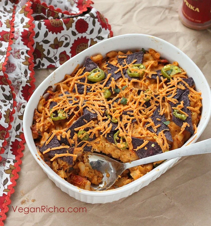 Baked vegan casserole topped with shredded cheese, sliced green peppers, and purple potato chips in a white oval dish, with a spoon in it, on a brown paper surface with a decorative cloth and a can in the background