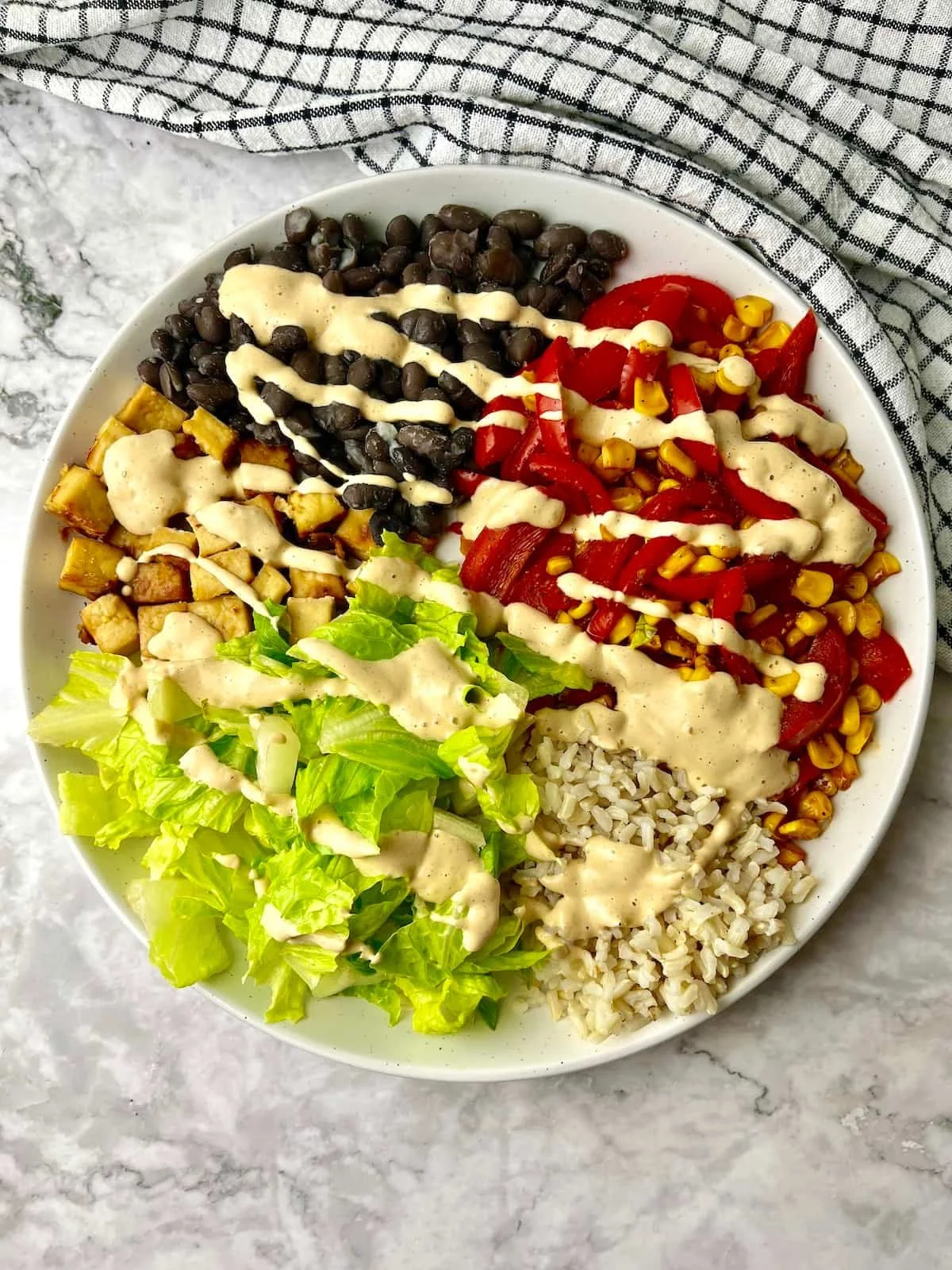 A white bowl filled with a salad and various toppings, including black beans, chopped red bell peppers, corn kernels, cooked rice, and chopped lettuce, all drizzled with a creamy dressing. The bowl is on a marble surface with a checkered cloth nearby.