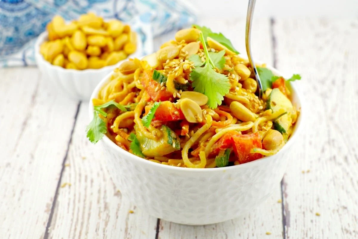 Bowl of Asian noodle salad with vegetables and peanuts on a white wooden surface, with a smaller bowl of peanuts in the background.