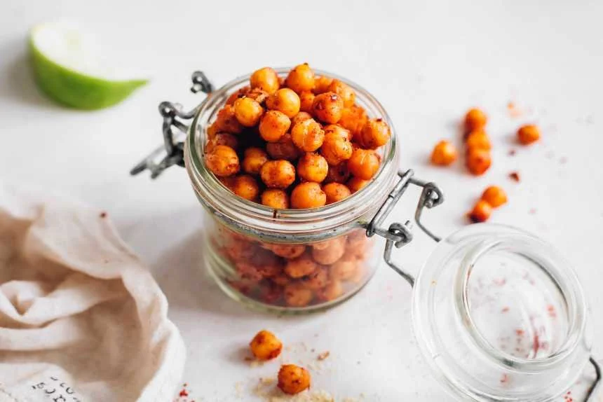A glass jar filled with spicy roasted chickpeas on a white surface with a lime wedge and a cloth napkin nearby.