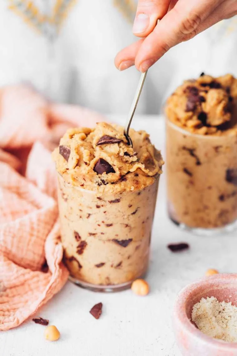 A spoon being used to scoop vegan cookie dough ice cream with chocolate chunks from a glass container, with another similar container in the background and a pink bowl of sprinkles nearby.