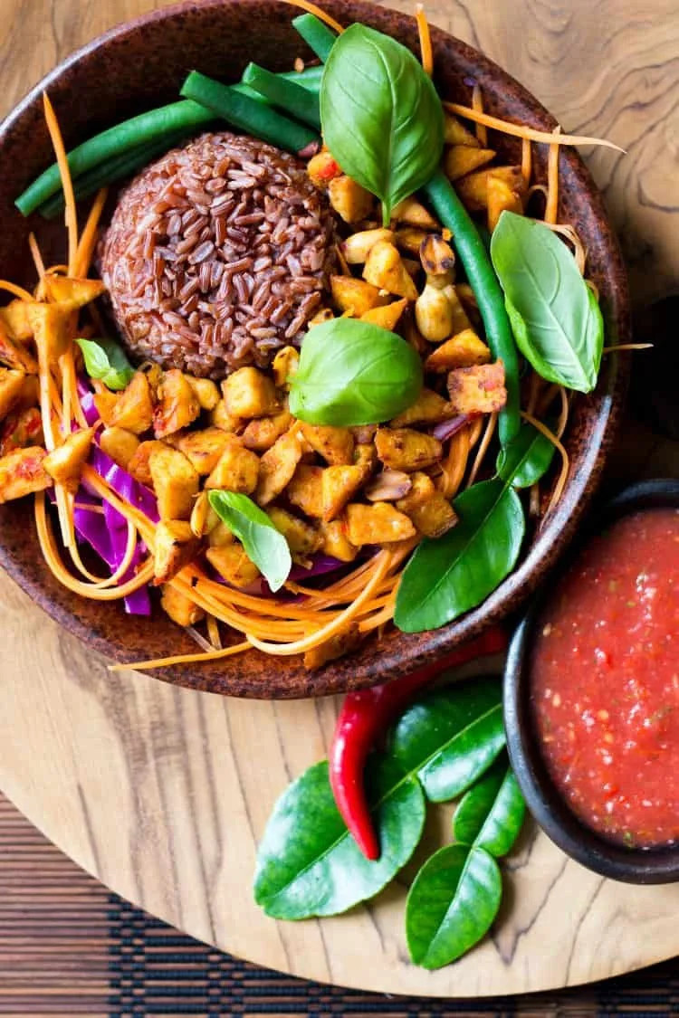 A bowl of Asian-inspired dish with brown rice, diced grilled chicken, green beans, purple cabbage, and fresh basil leaves, served with a side of red chili pepper and green lime on a wooden board.