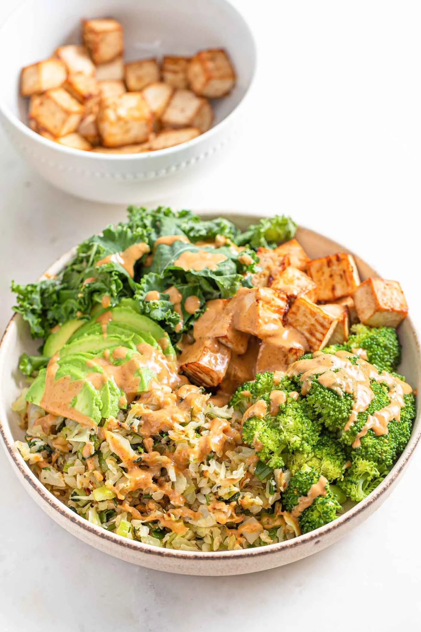 Tofu Power Bowl salad with chopped greens, sliced avocado, chopped cabbage, and broccoli, all topped with a creamy dressing. In the background, a smaller bowl with diced cooked tofu or meat.