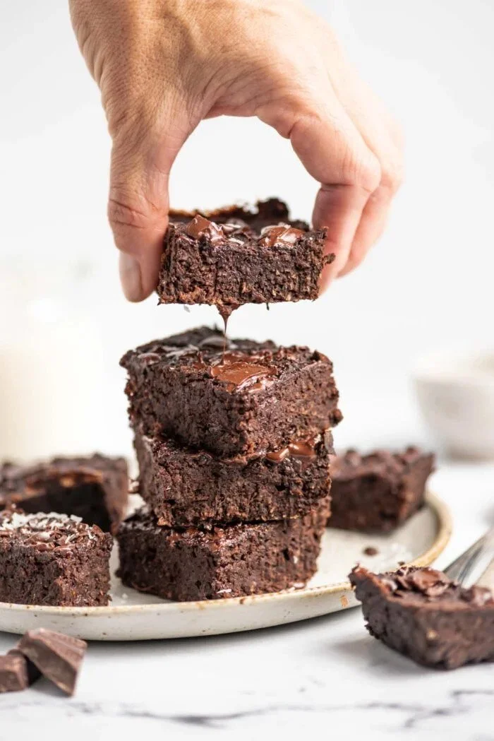 A hand stacking three chocolate brownies on a white plate, with melted chocolate being dripped onto the top brownie.