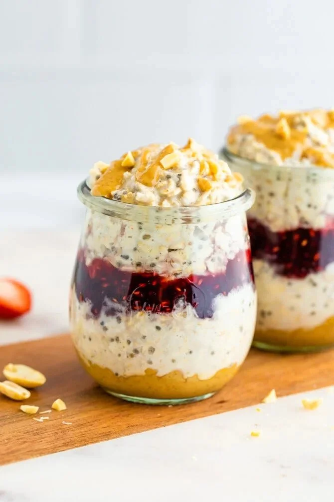 Layered dessert in a jar with peanut butter, whipped cream, berry jam, and cookie crumbs topped with chopped nuts.