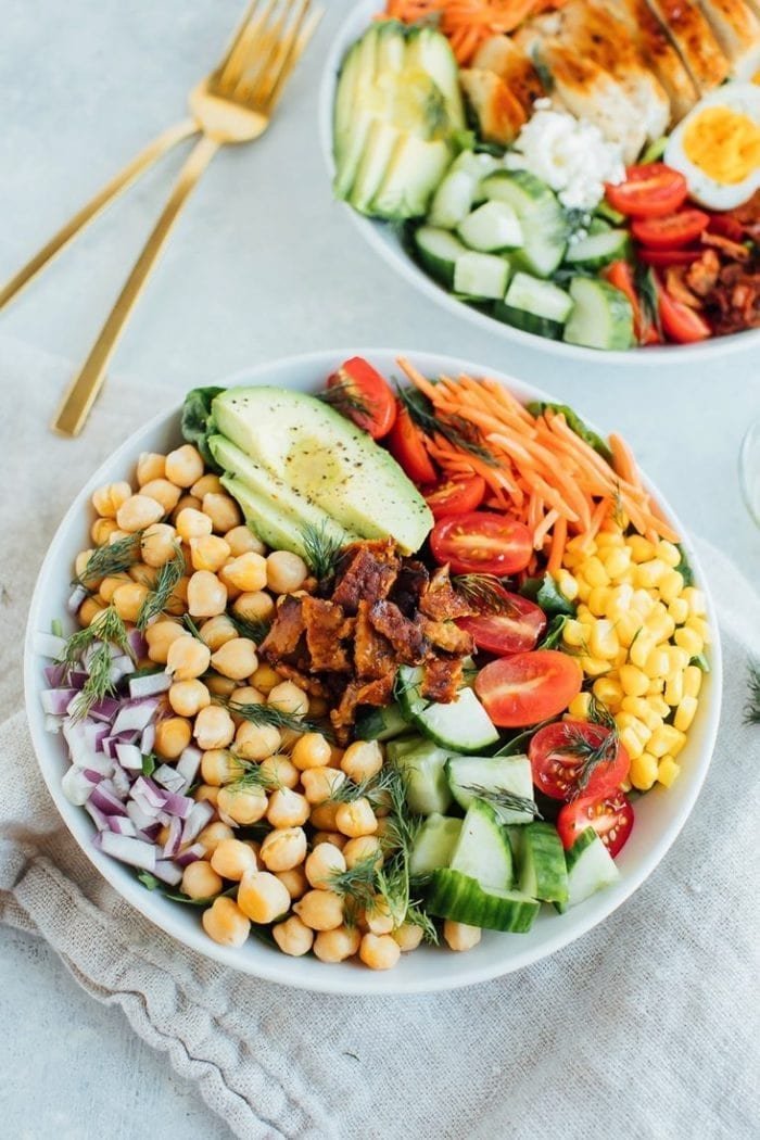 A bowl of colorful chopped vegetables and chickpeas, including cherry tomatoes, cucumber, onion, carrots, avocado, and herbs, placed on a white tablecloth with a gold fork nearby.