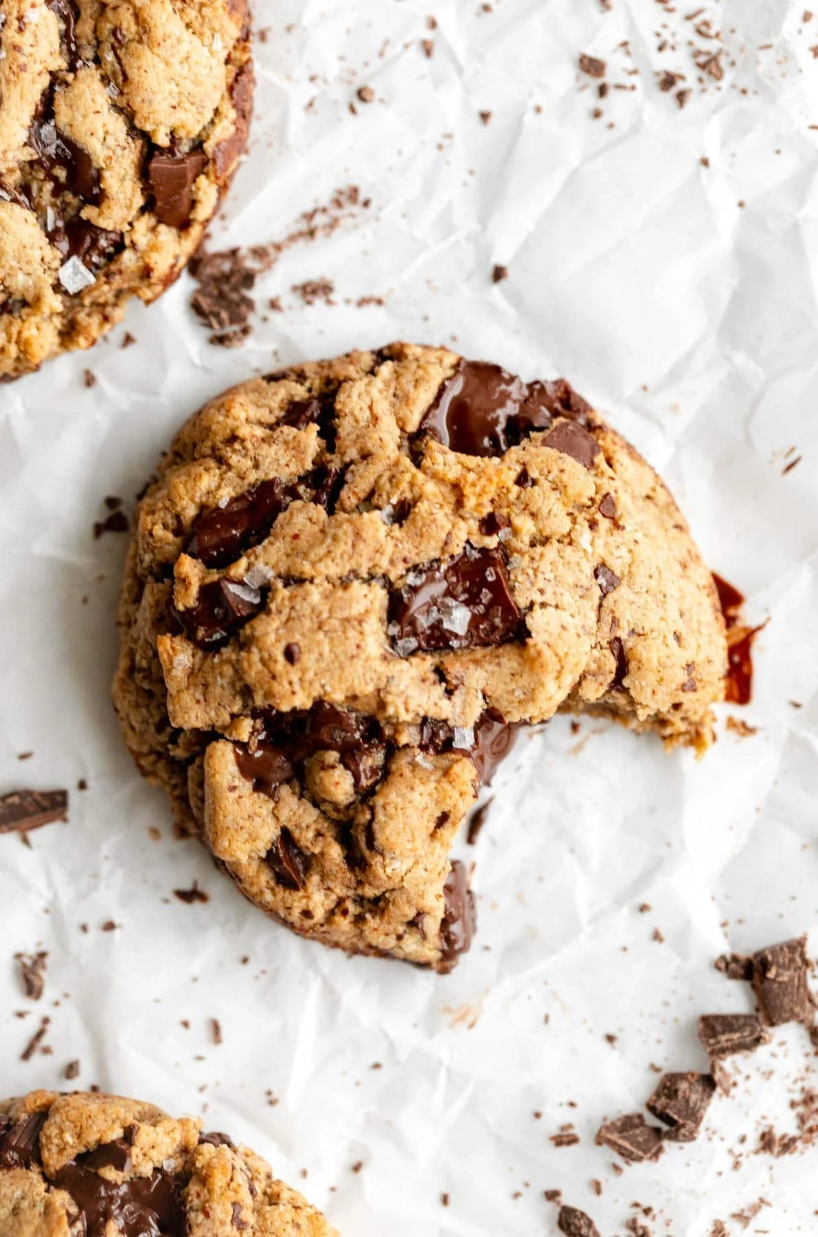 Close-up of a chocolate chip cookie with a bite taken out, resting on crumpled white paper with chocolate shavings nearby.