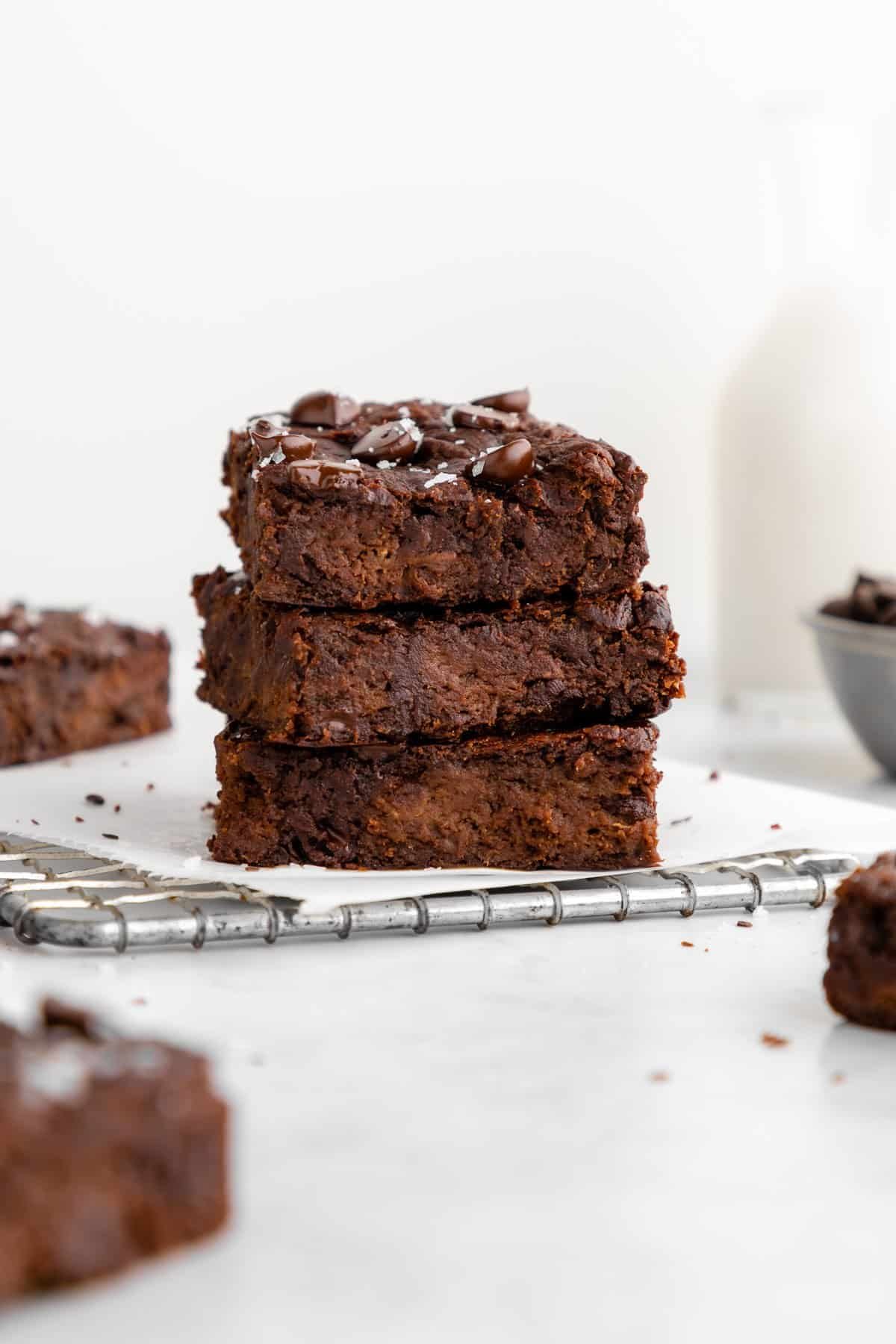 Stack of three chocolate brownies topped with chocolate chips on parchment paper, with a bowl of more brownies in the background.