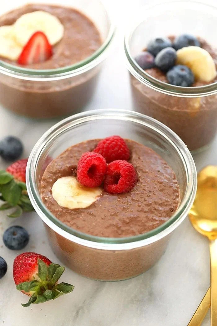 Three glass jars of chocolate mousse topped with assorted berries, including raspberries, blueberries, and banana slices, with fresh strawberries and blueberries scattered around.