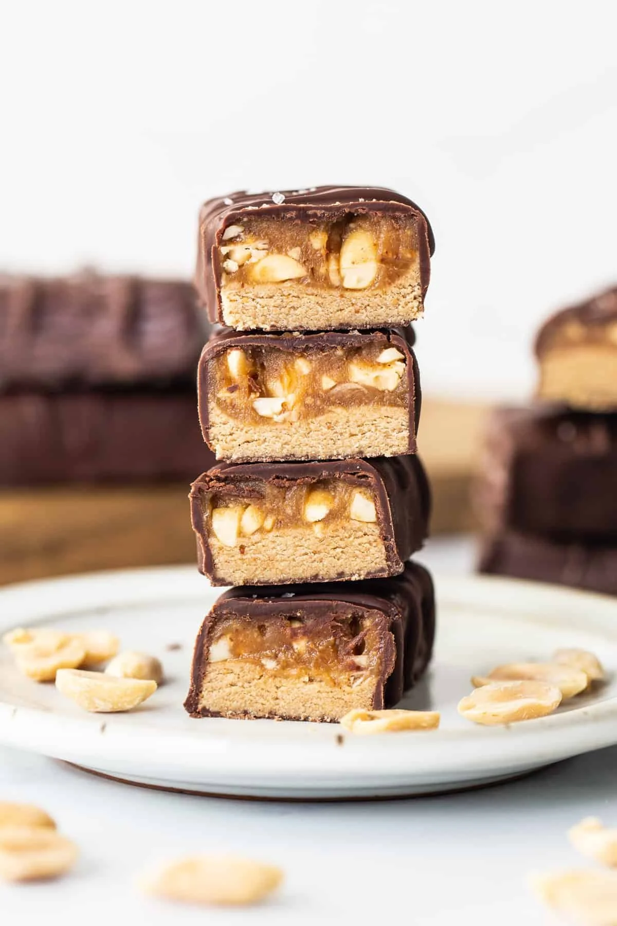 Stacked caramel and peanut chocolate candies on a white plate with scattered peanuts, with more candies in the background.