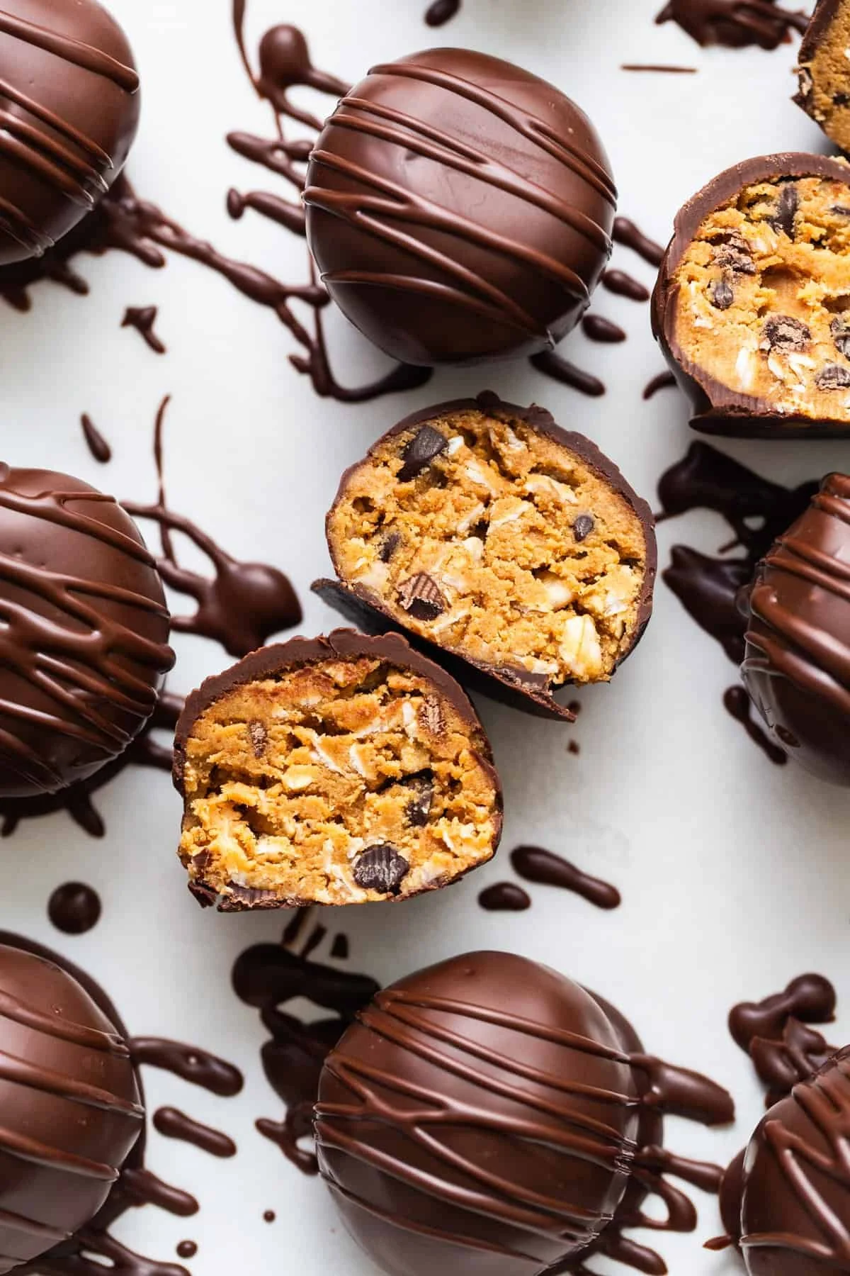 Chocolate-covered cookie dough truffles with drizzled chocolate, some cut in half to show the cookie dough and chocolate chip filling inside, on a white surface with chocolate drizzles and dollops.