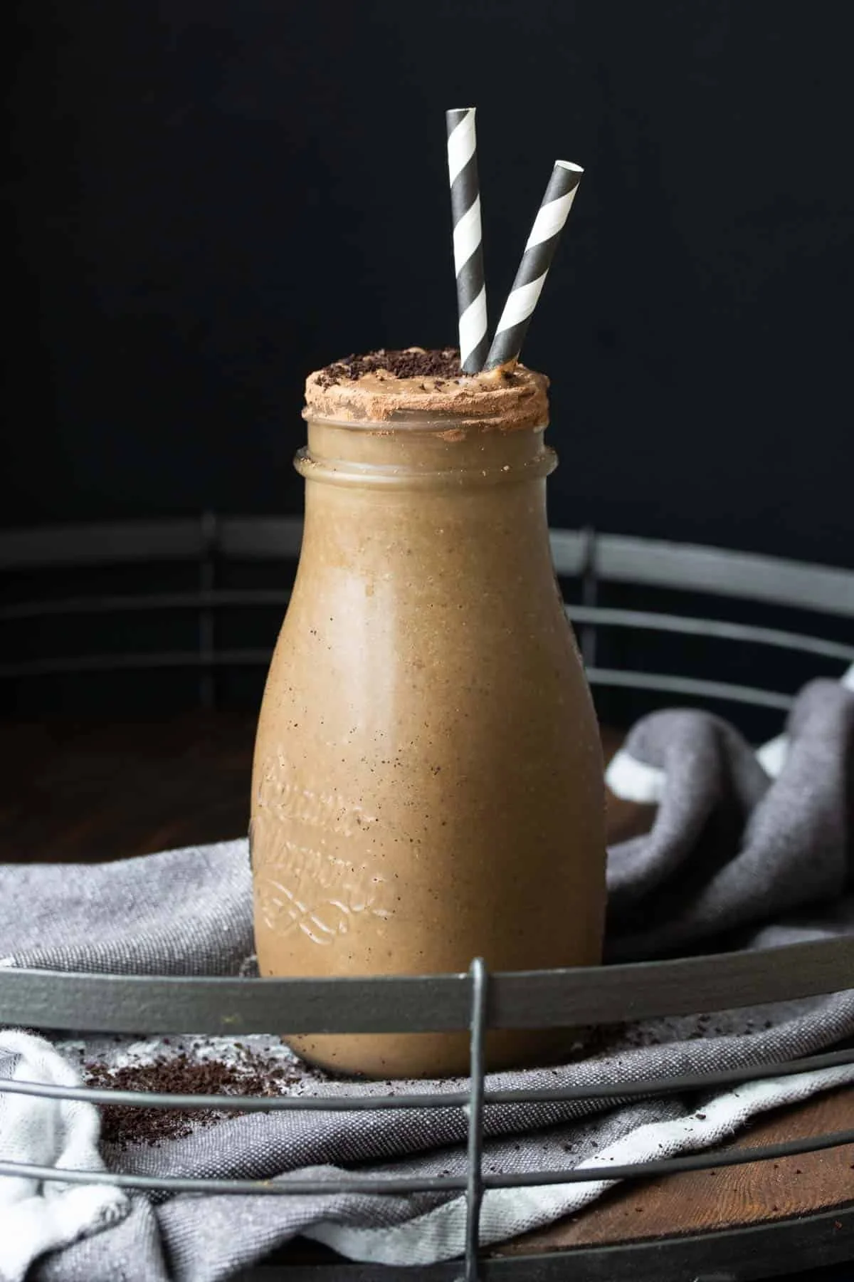 A glass bottle filled with a creamy vegan chocolate shake, topped with crushed cookies and two black-and-white striped paper straws, placed on a cloth with a dark background.