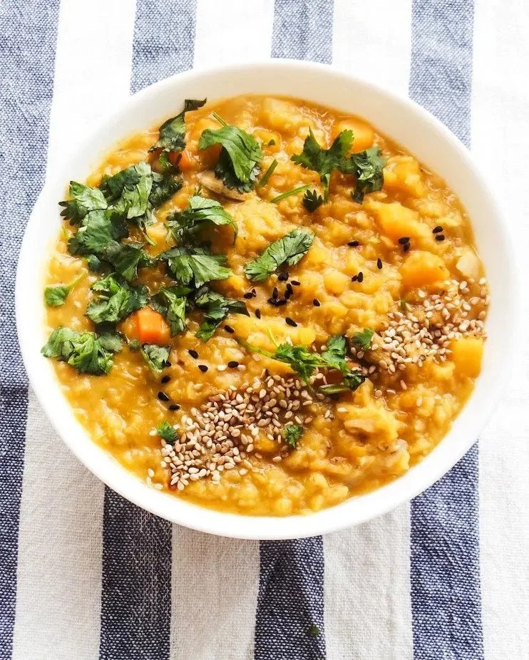A bowl of lentil soup topped with chopped cilantro, sesame seeds, and black seeds, placed on a striped cloth background.
