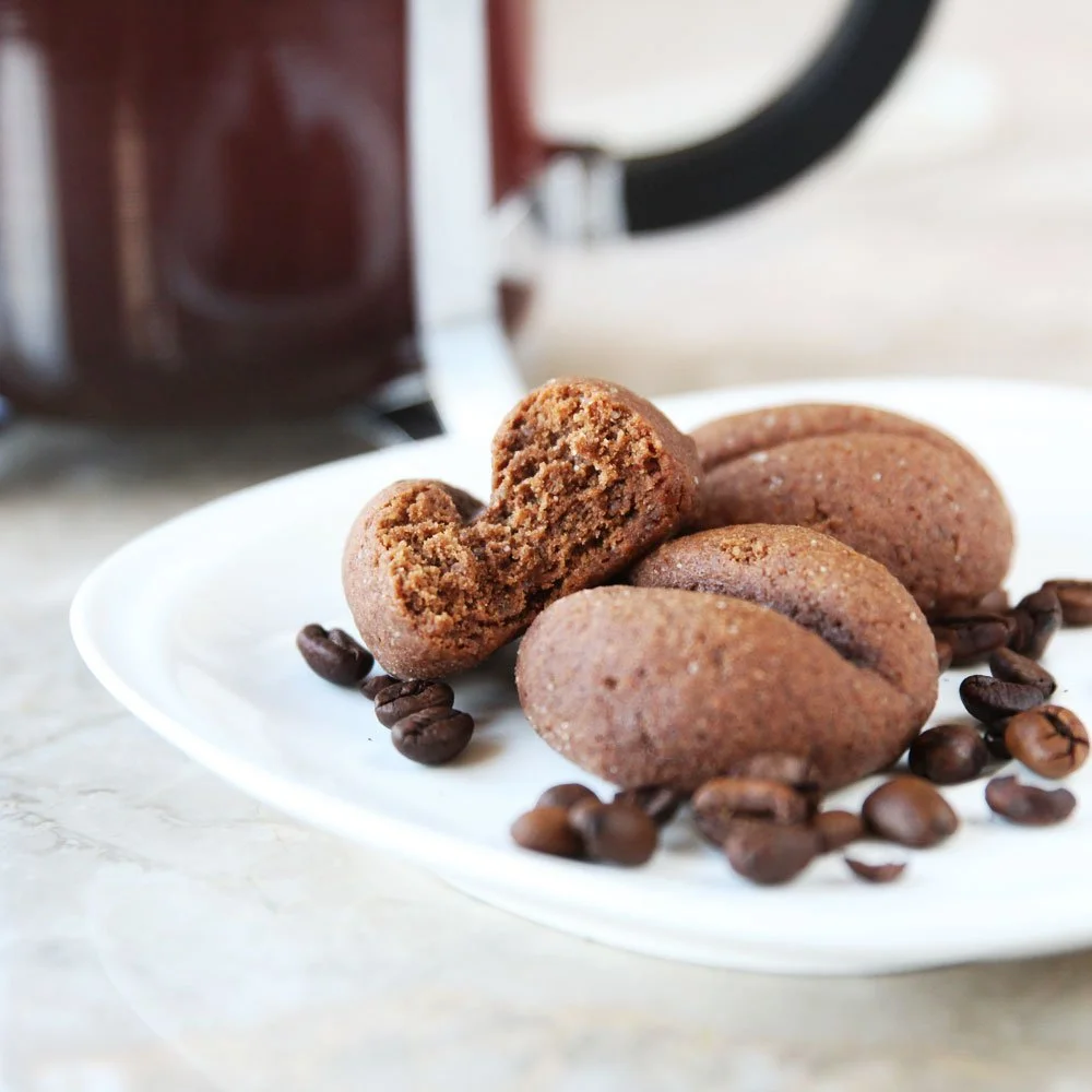 Chocolate cookies on a white plate with coffee beans scattered around, with a coffee pot in the background.