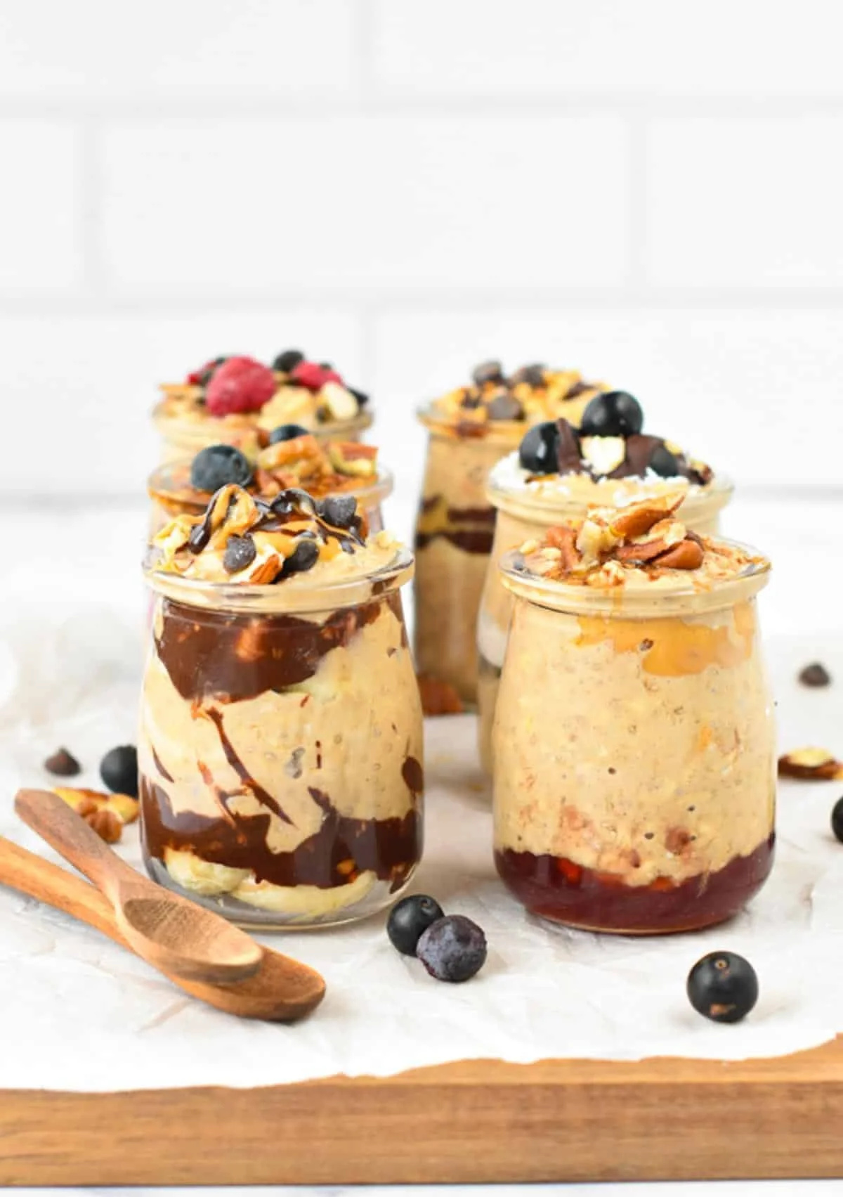 Six glass jars filled with layered vegan dessert topped with blueberries, nuts, chocolate, and caramel, set on a wooden board with scattered blueberries and a wooden spoon.