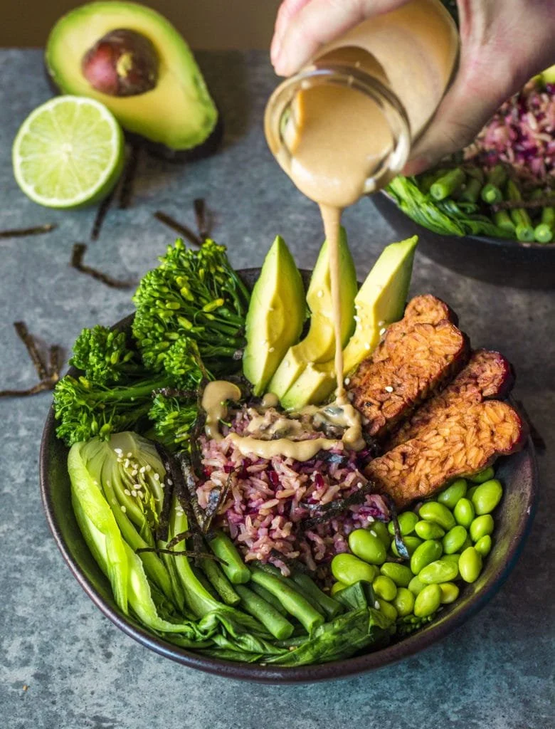 A bowl of vegetarian food with rice, avocado slices, broccoli, cucumber, edamame, crispy tempeh, and a drizzle of peanut sauce, with lime and avocado in the background.