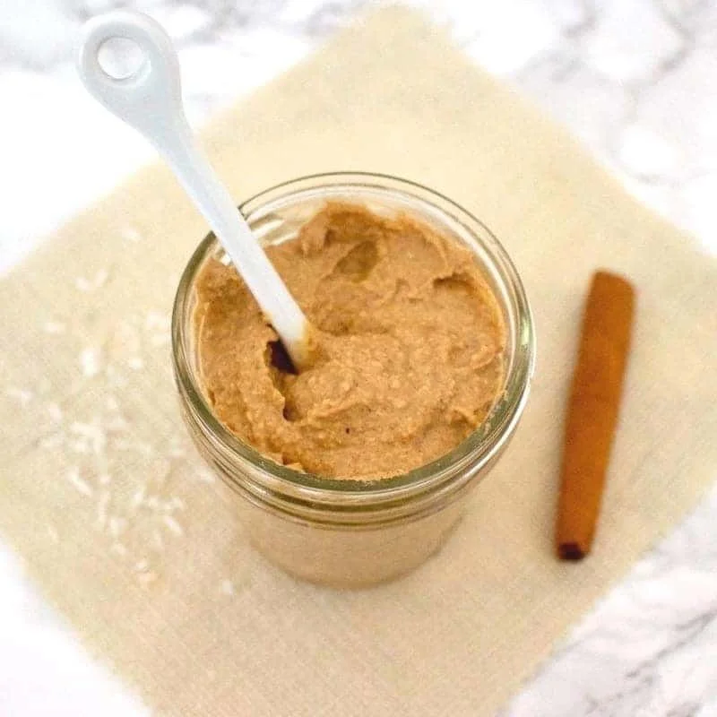 A glass jar of cinnamon-spiced peanut butter with a white spoon inside, placed on a white napkin with a cinnamon stick beside it.