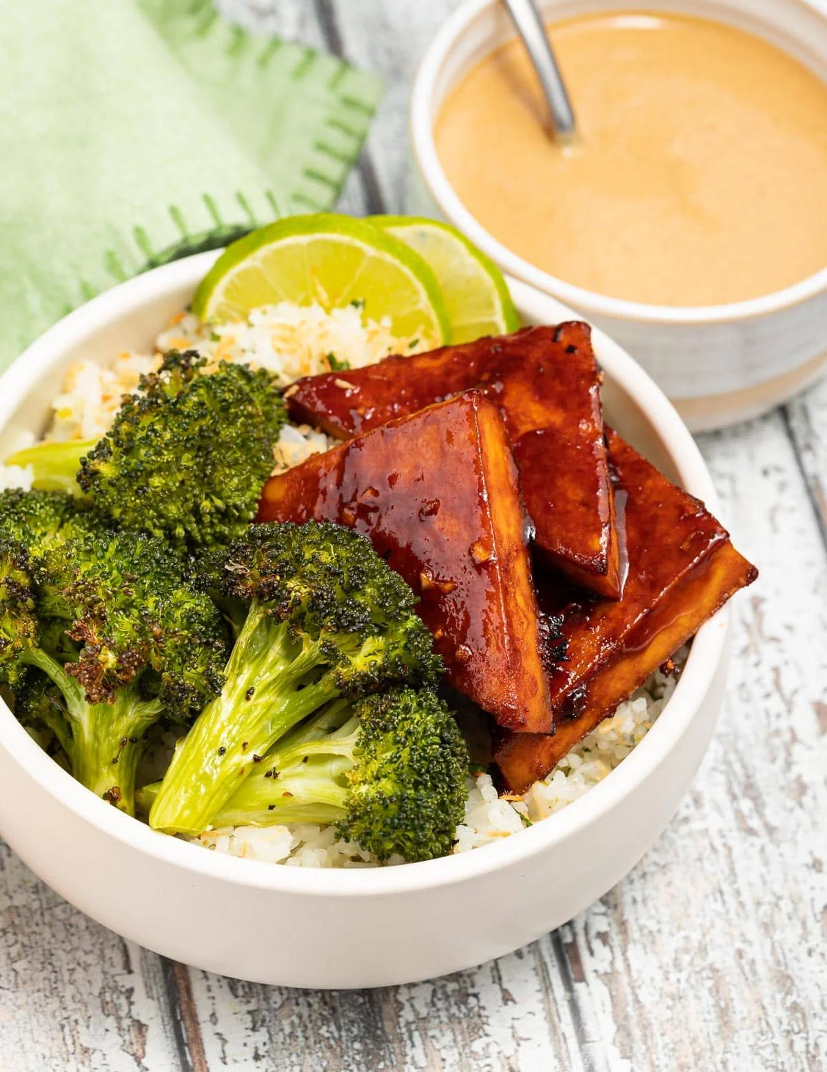 Bowl of rice topped with barbecue tofu, broccoli, and lime slices, served with a cup of tea or coffee on a wooden table.