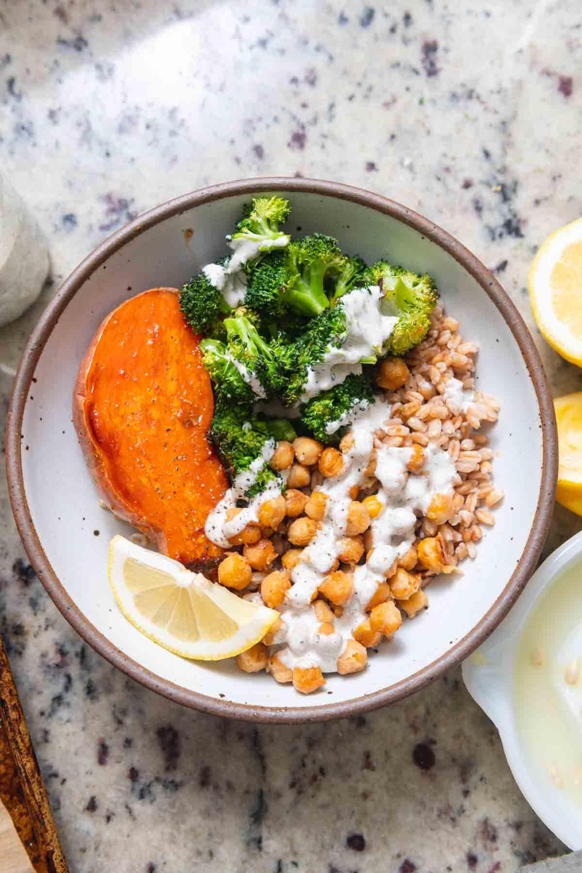 A bowl containing roasted sweet potato, steamed broccoli topped with white sauce, cooked chickpeas with white sauce, and a lemon wedge on a speckled granite surface.