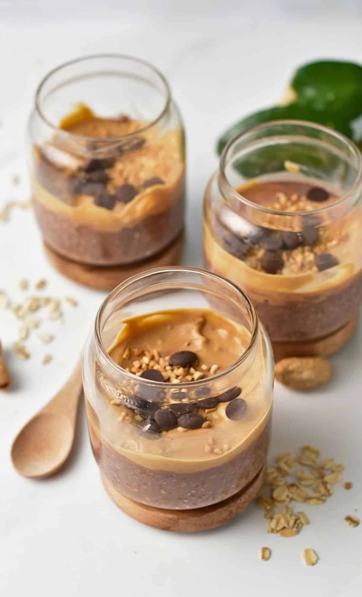 Three glasses of layered peanut butter mousse topped with chocolate chips and chopped nuts, with a wooden spoon and scattered oats on a white surface.