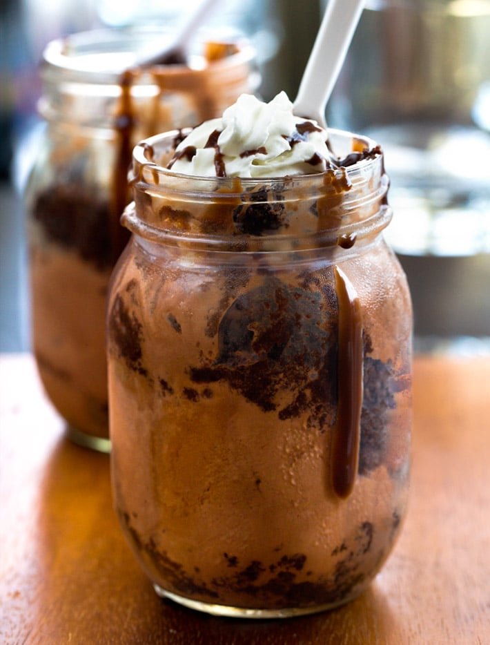 Close-up of a chocolate milkshake in a glass jar topped with whipped cream and chocolate syrup, with another similar milkshake in the background.