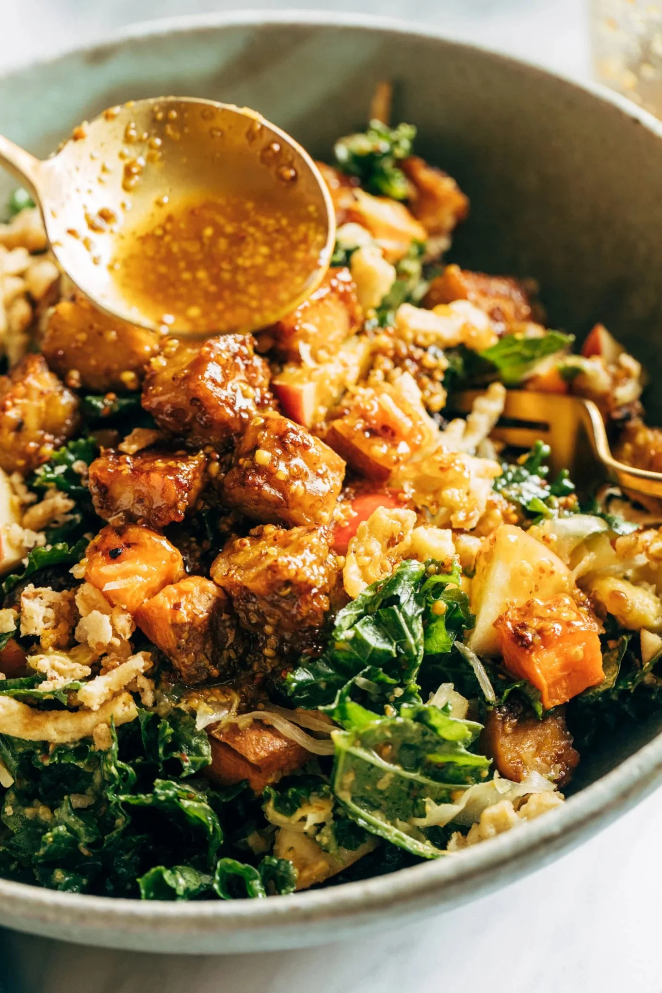 Close-up of a salad in a bowl with chunks of roasted vegetables, leafy greens, shredded cheese, and a spoon pouring salad dressing.
