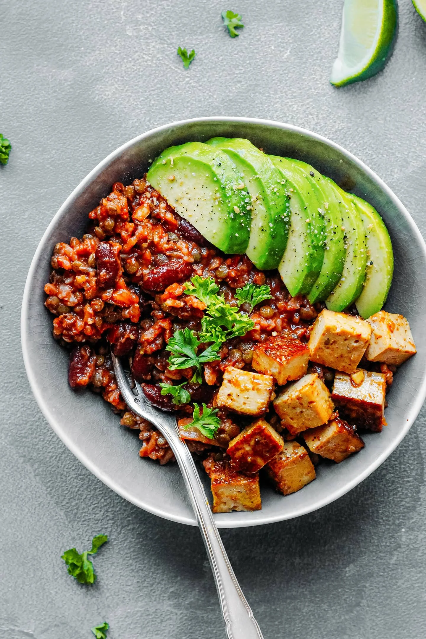 Tofu power bowl with riece, sliced avocado, and a sprig of cilantro on top, with a lime wedge and scattered herbs nearby on a gray surface.