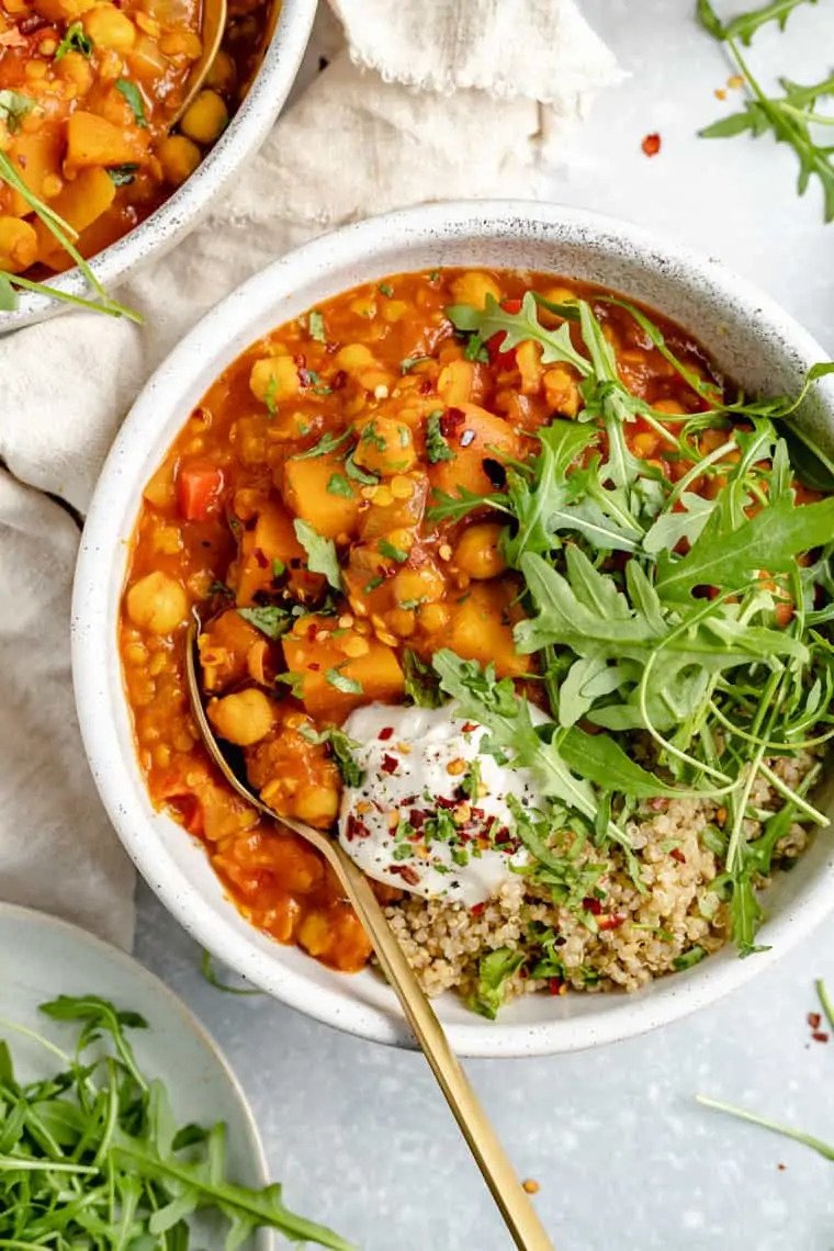 A bowl of chickpea vegetable curry with fresh arugula, quinoa, yogurt, and red pepper flakes