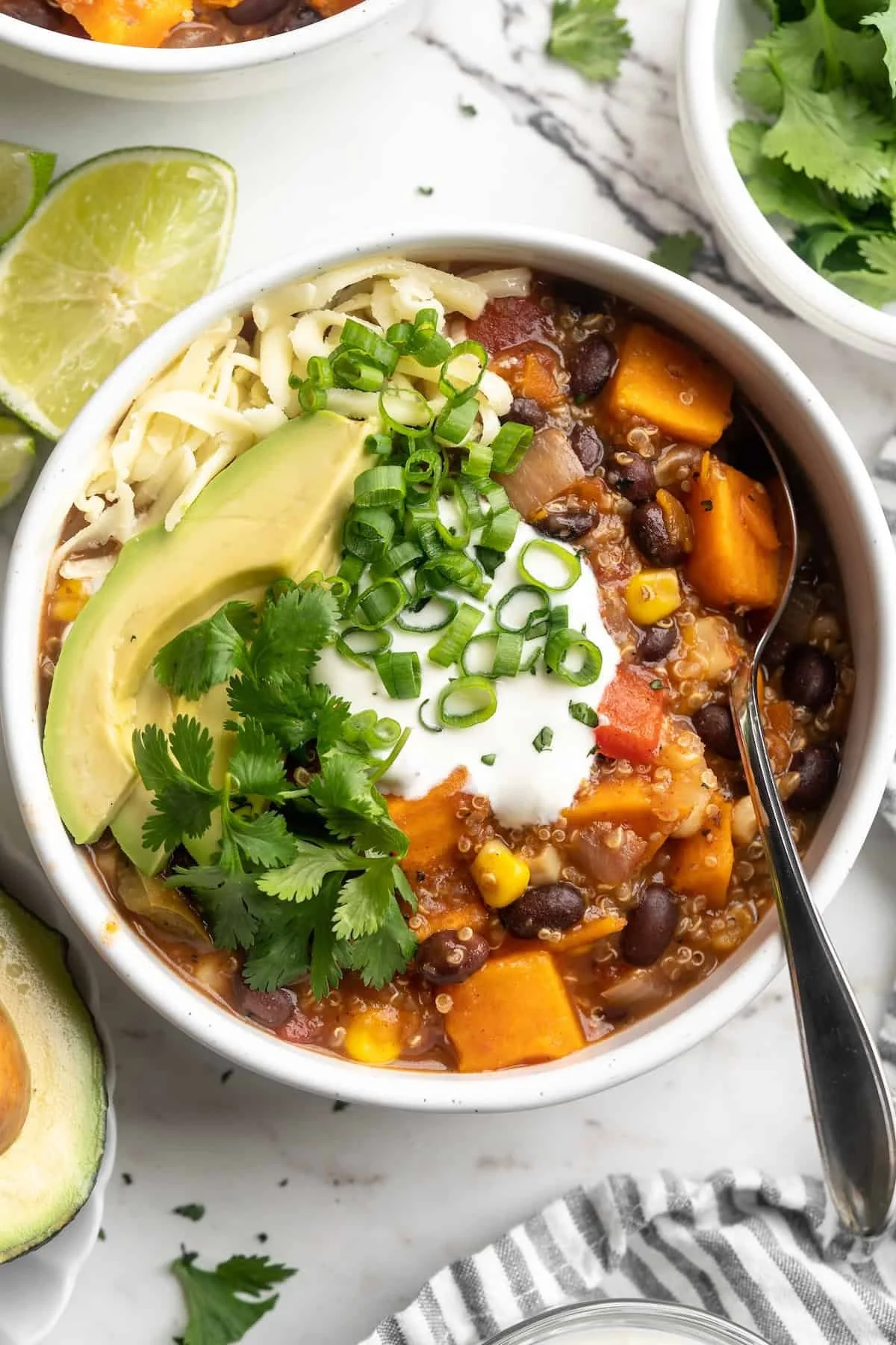 A bowl of chili topped with diced green onions, sour cream, cilantro, shredded cheese, avocado slices, and a lime wedge.