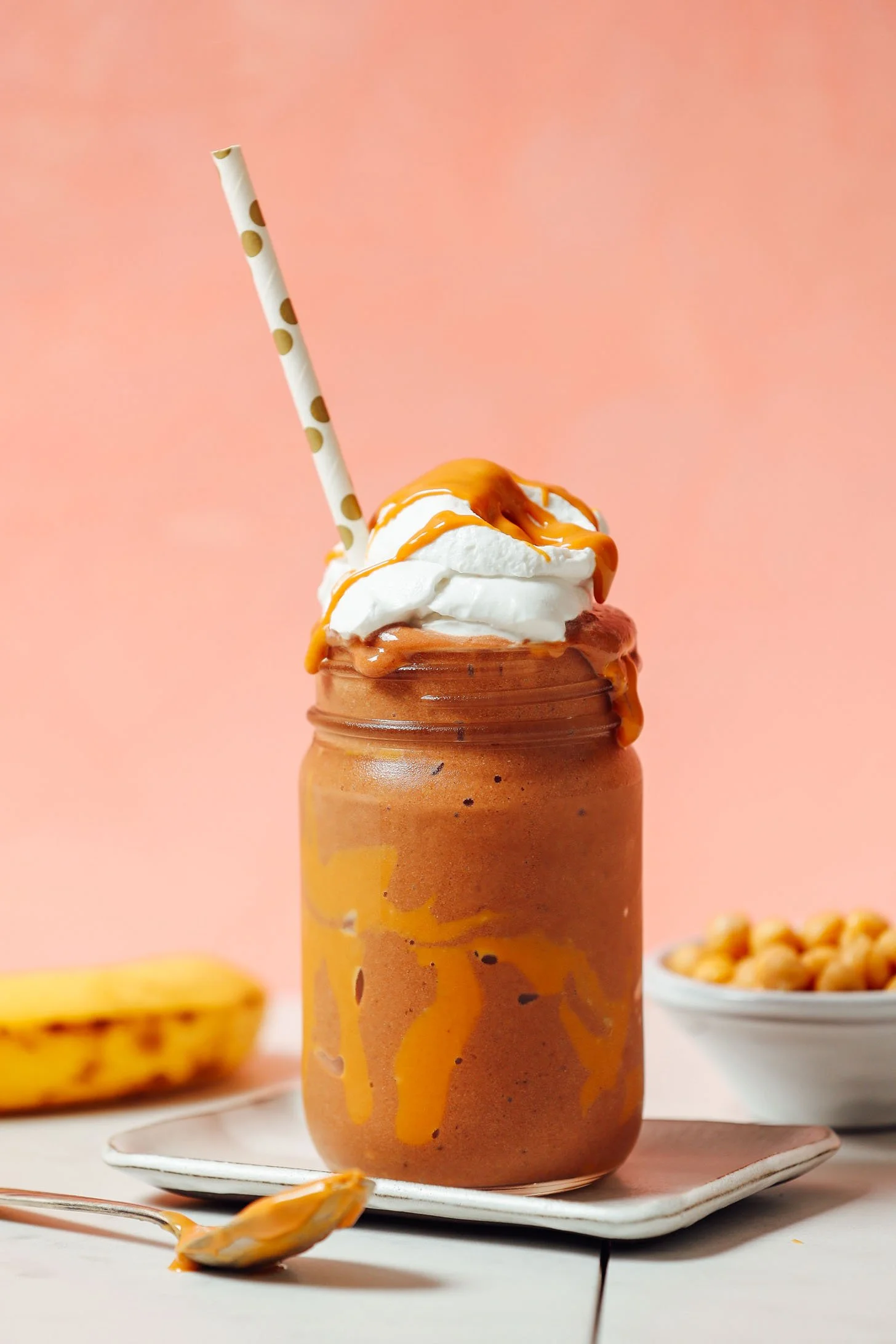 A vegan caramel frappuccino in a glass jar topped with whipped cream and caramel drizzle, with a polka-dotted straw, placed on a white plate with a spoon, a banana, and a bowl of cereal in the background against a pink wall.
