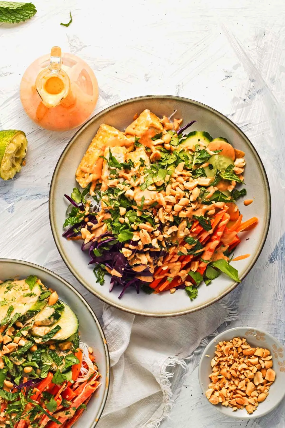 A bowl of mixed vegetable salad with shredded carrots, cucumbers, purple cabbage, chopped peanuts, and fresh herbs, with a small bowl of chopped peanuts and a small pitcher of dressing on the side.