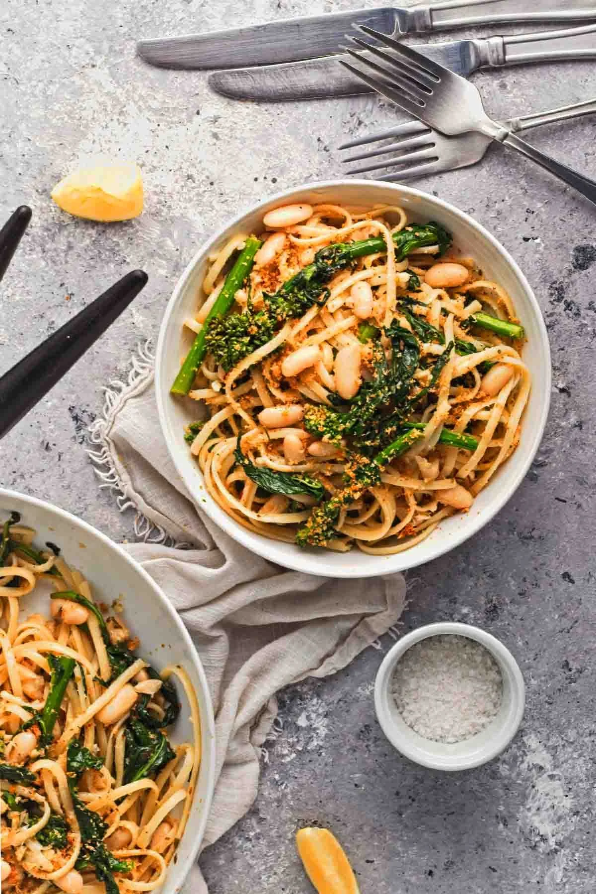 A bowl of spaghetti with beans, green vegetables, and sprinkled seasoning, on a textured surface with utensils, lemon wedge, and salt nearby.