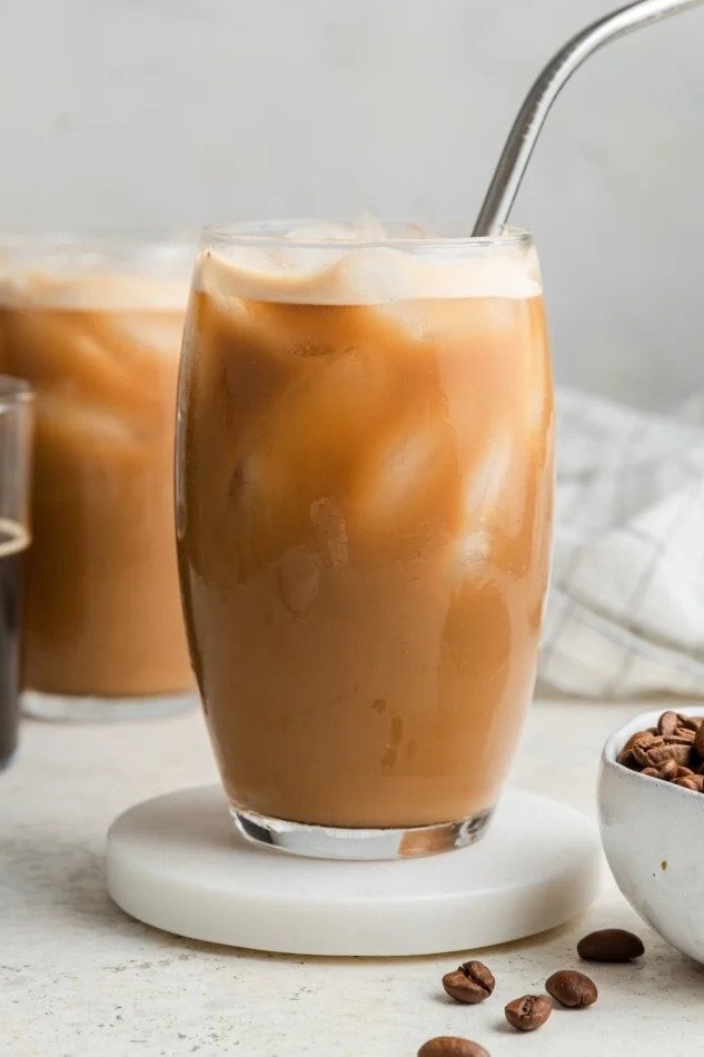 A tall glass of iced coffee with a metal straw, placed on a white circular coaster. There is a bowl of coffee beans next to it and a blurred second glass of iced coffee in the background.