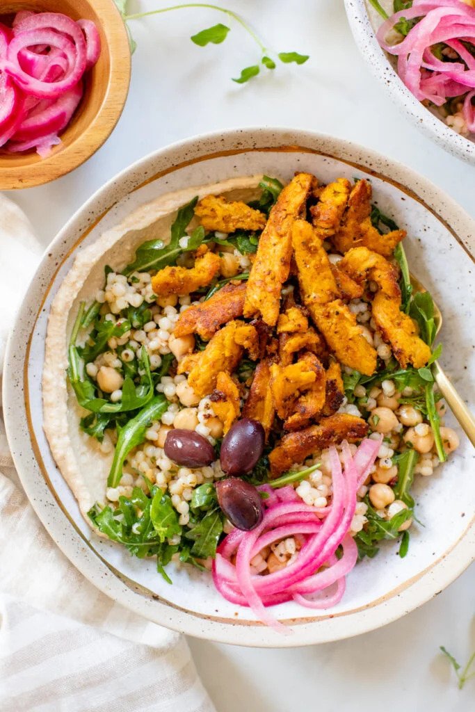A bowl of vegan salad containing grilled plant-based chicken strips, arugula, couscous, Kalamata olives, and pickled onions.