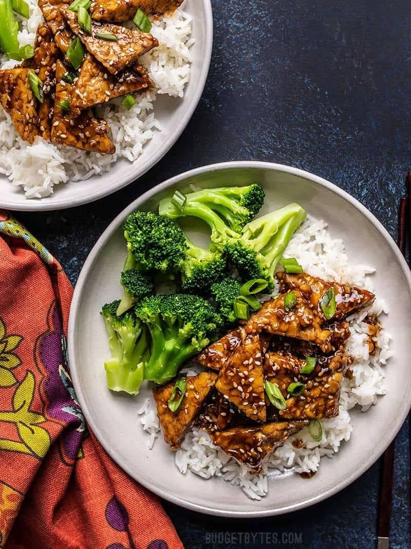 Plate of white rice topped with teriyaki chicken garnished with chopped green onions, with steamed broccoli on the side, on a dark surface with colorful cloth.