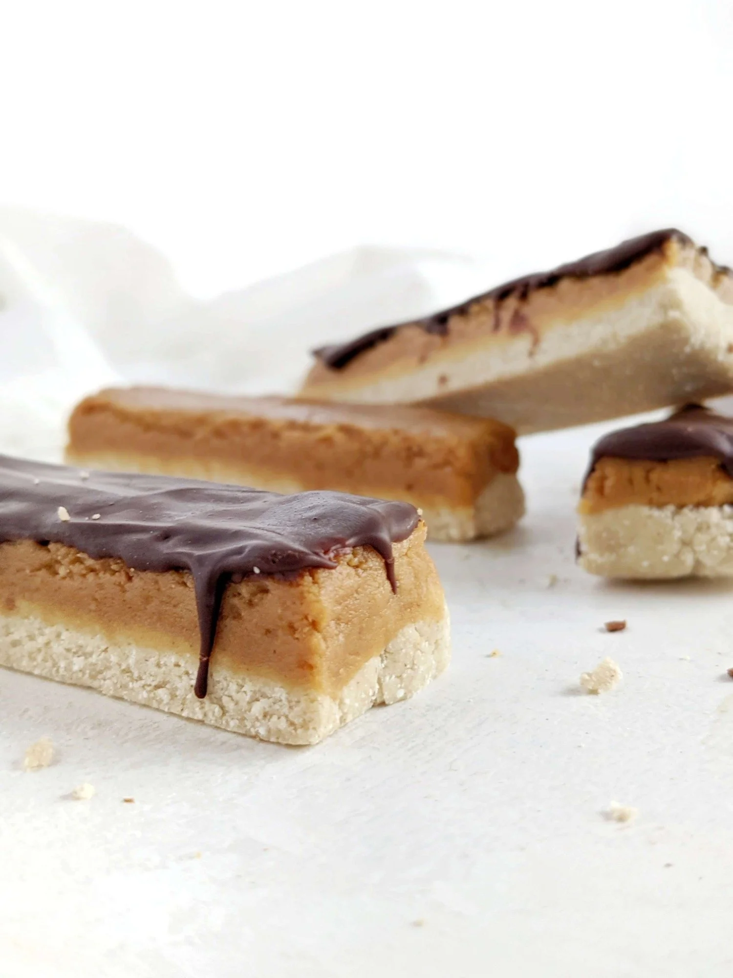 Close-up of three rectangular cookies with a layer of chocolate on top, arranged on a white surface with a crumpled white paper background.
