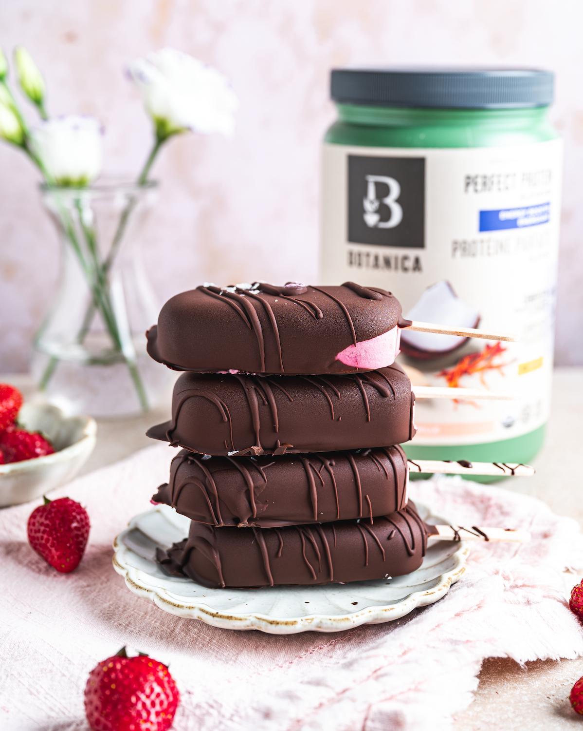 Four chocolate-coated ice cream bars with pink filling on a decorative white plate, surrounded by strawberries, with a container of plant-based protein powder and a glass vase with white flowers in the background.