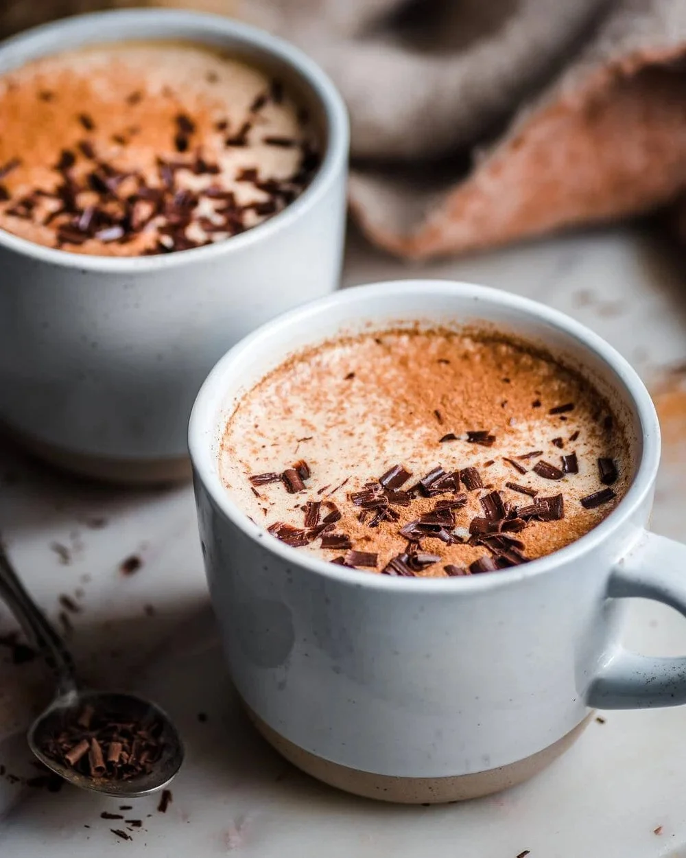 Two mugs of frothy vegan hot chocolate topped with chocolate shavings on a marble surface with a spoon of chocolate shavings beside.
