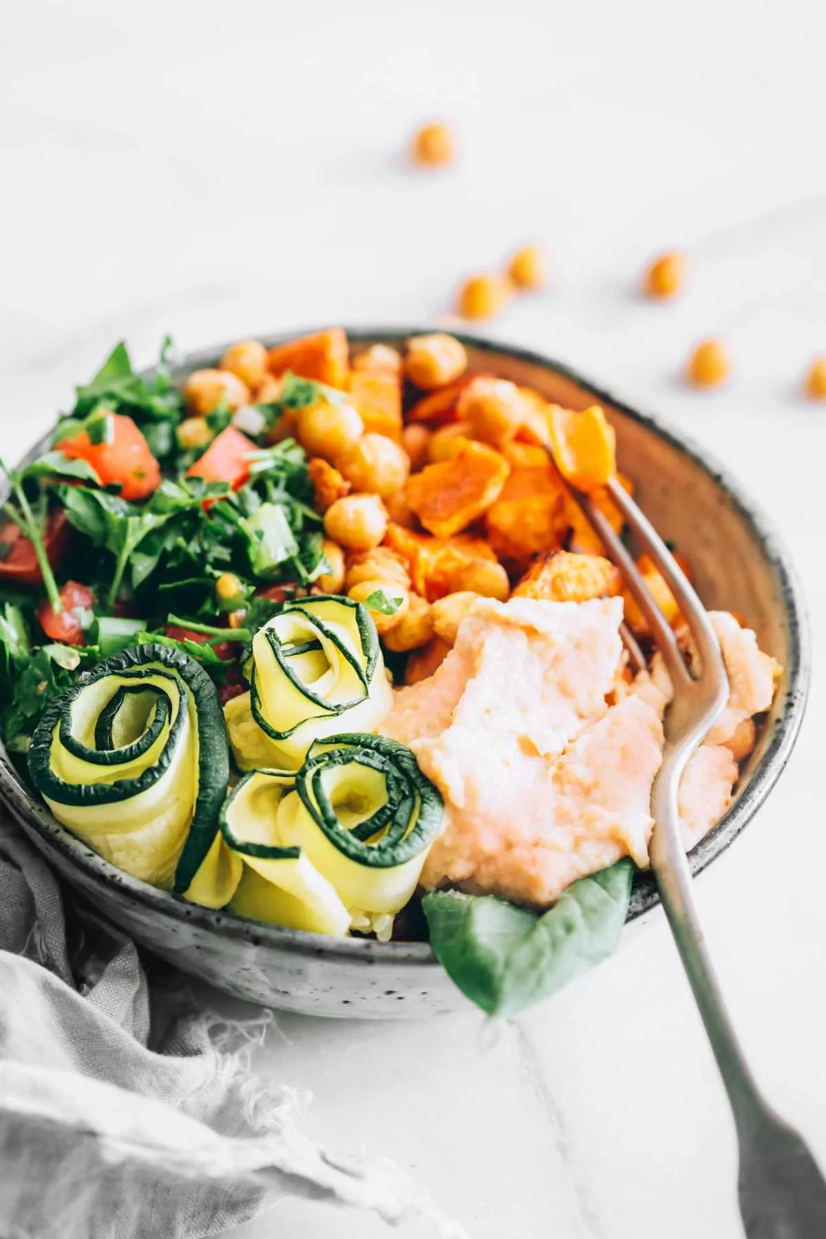A bowl of assorted vegetables including curly zucchini slices, chickpeas, shredded carrots, leafy greens, and mashed potatoes on a white surface.