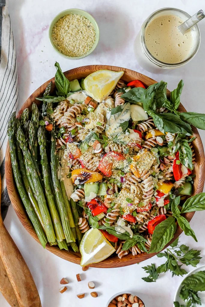 A large salad with whole wheat pasta, cherry tomatoes, avocado, basil leaves, and grated cheese in a wooden bowl, with a side of green beans, lemon wedges, a glass of creamy dressing, and a small bowl of grated cheese on a white surface.